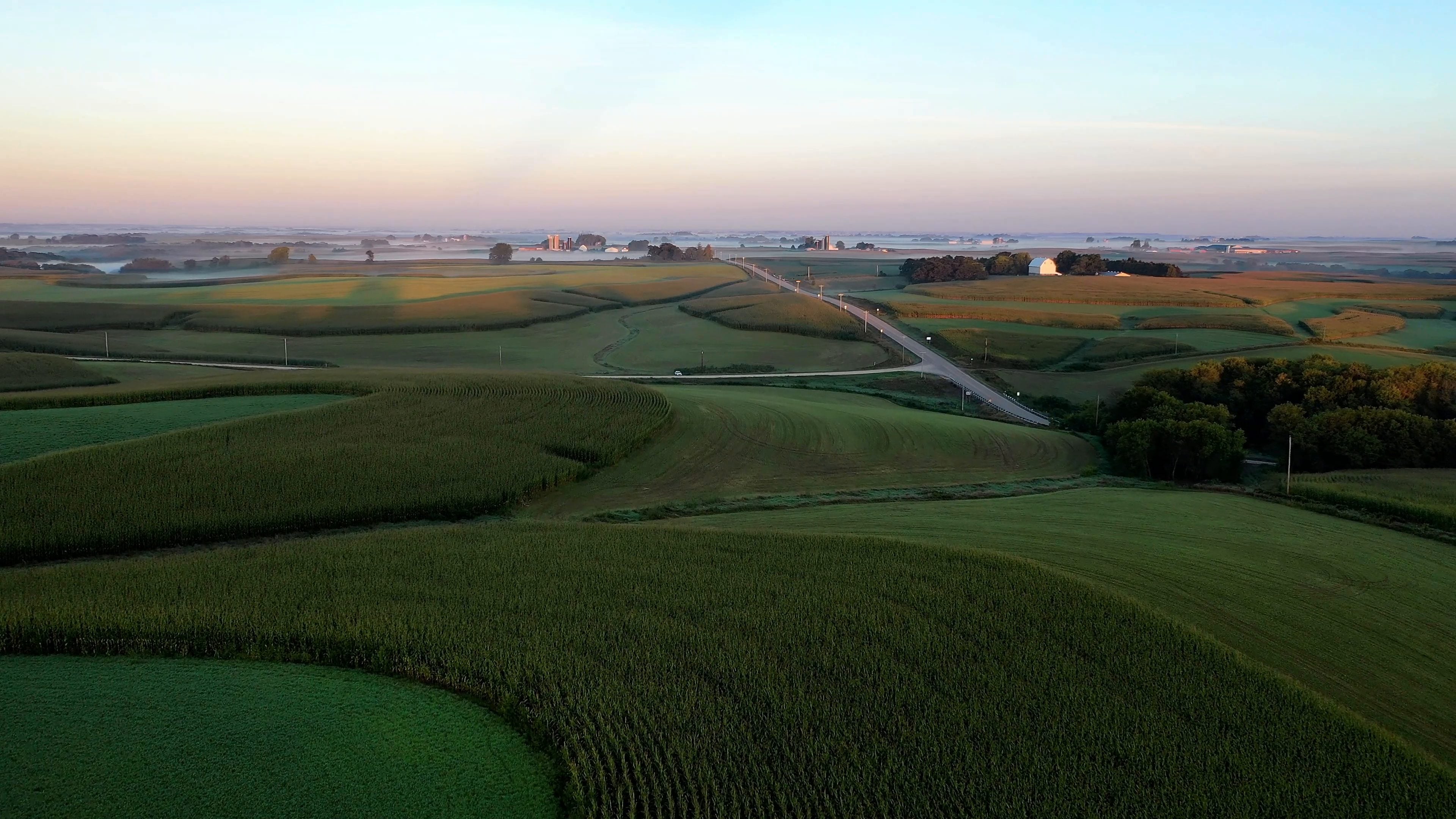 Aerial view of rolling farmland at sunrise, with crop strips planted along the contours, a rural road, a white barn and silos in the distance, and low mist on the horizon.