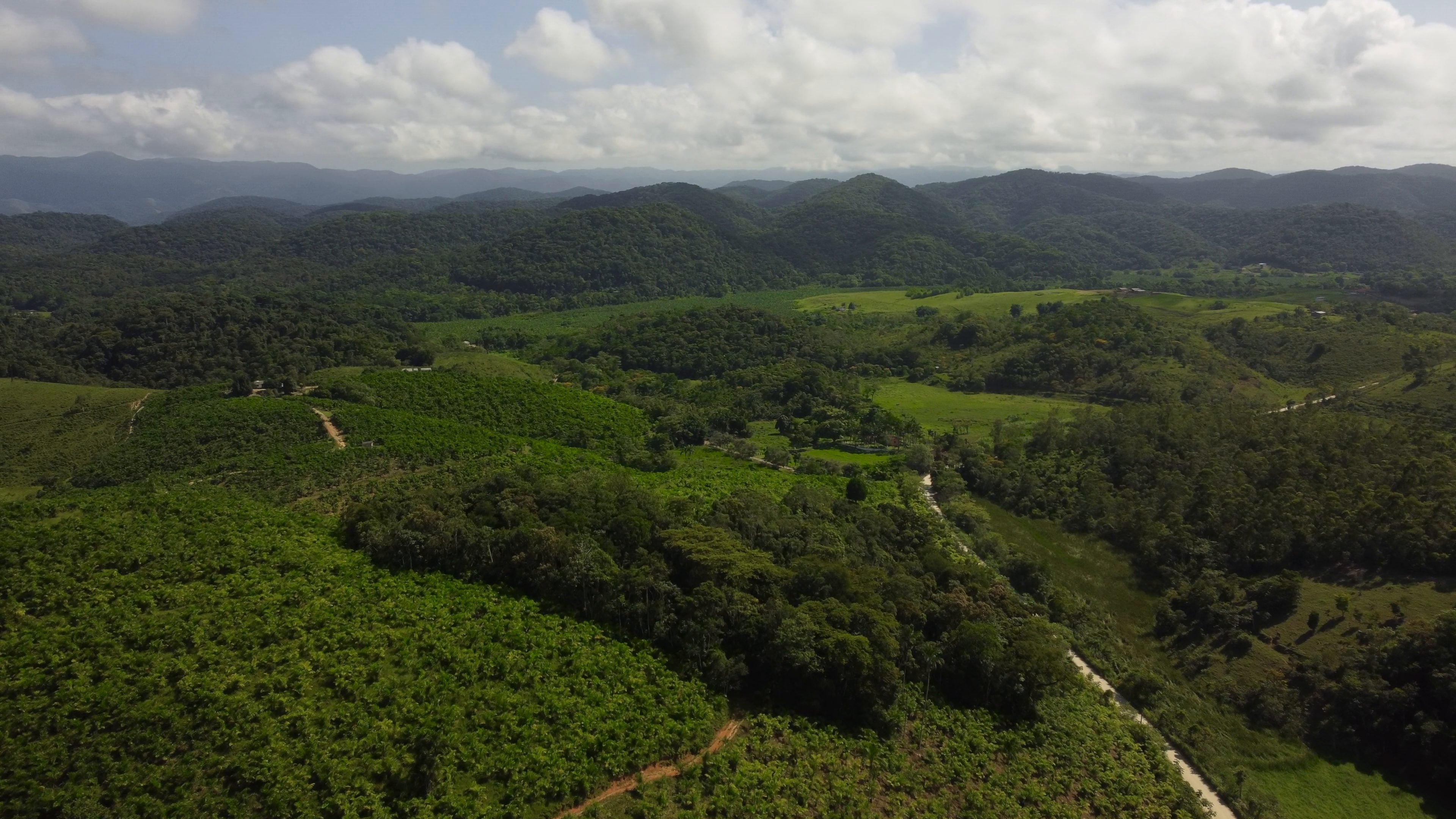 Aerial view of hills in Brazil, with crops alongside patches of forest, dirt roads and pastures, and mountain ranges under clouds beyond.